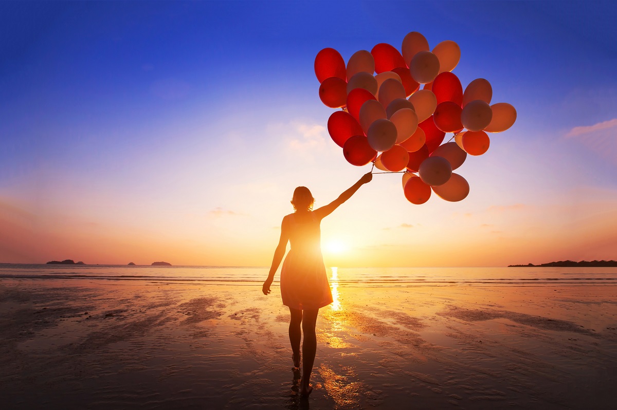 A woman holding a bunch of red balloons in front of a sunset on a beach